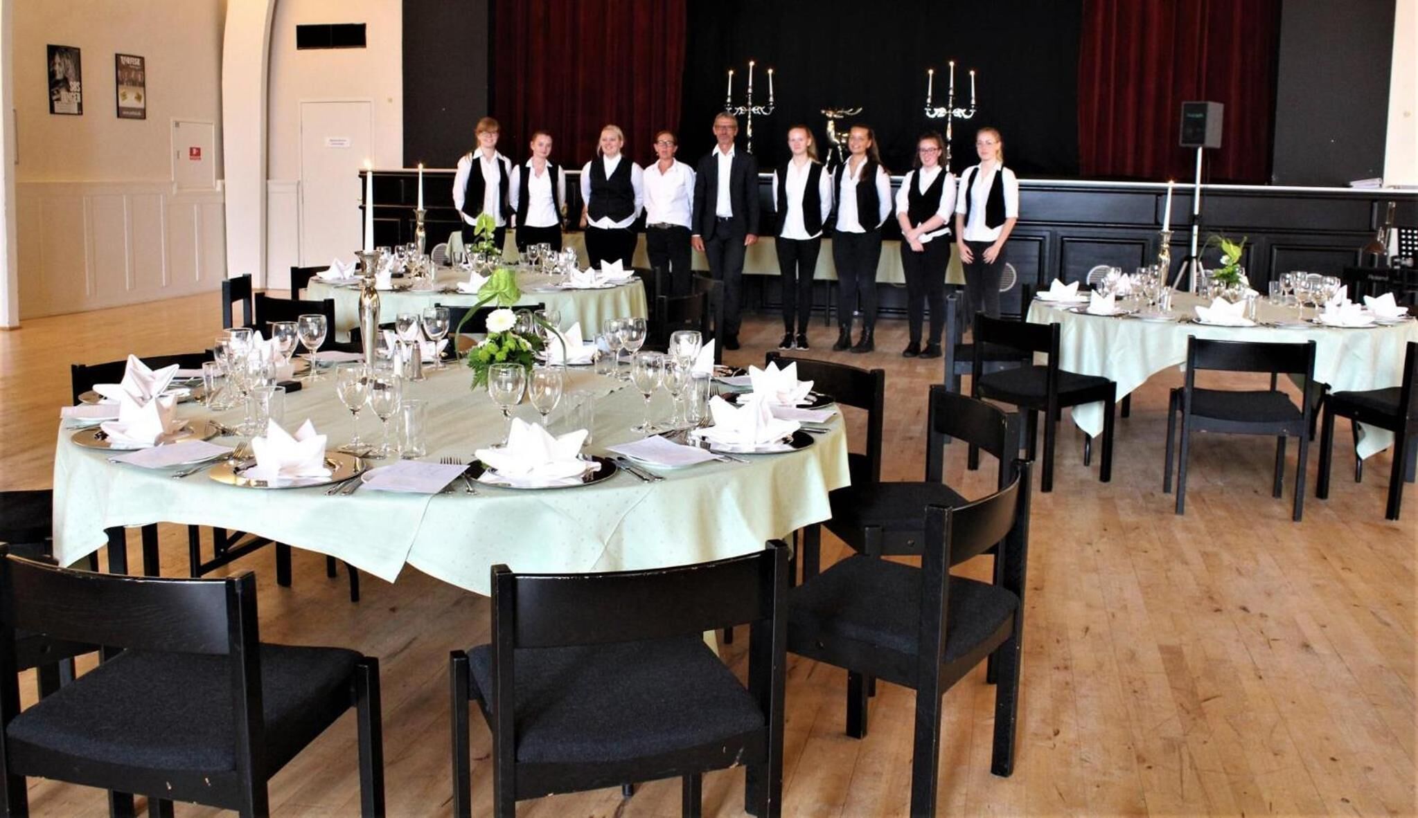 Un comedor formal con mesas redondas dispuestas para una comida, sillas negras y un escenario con velas al fondo.