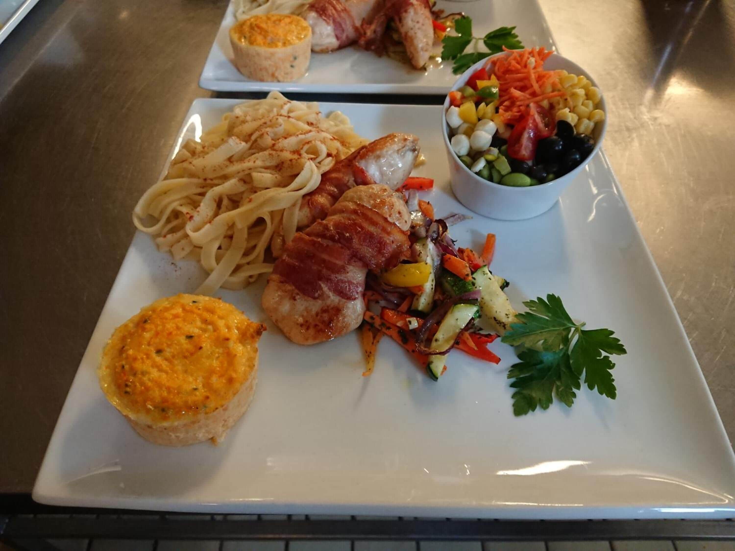 A plated meal with pasta, a piece of chicken with bacon, a muffin, and a salad.