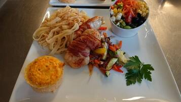 A plated meal with pasta, a piece of chicken with bacon, a muffin, and a salad.