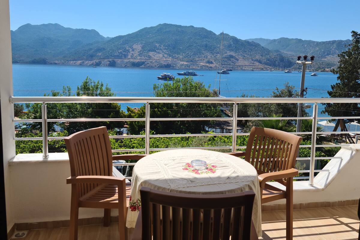 A balcony with a table and two chairs, overlooking a body of water with boats and mountains in the background.