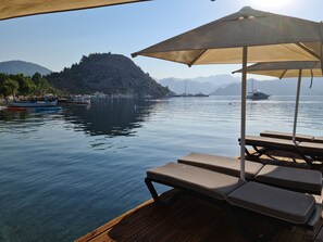 A wooden deck with lounge chairs and a canopy, overlooking calm water with boats and a mountainous backdrop. - Akkum Gezer Motel (Marmaris)