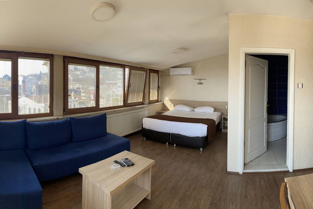 Habitación de hotel con una cama grande, un sofá, un televisor y vistas de la ciudad a través de amplios ventanales.