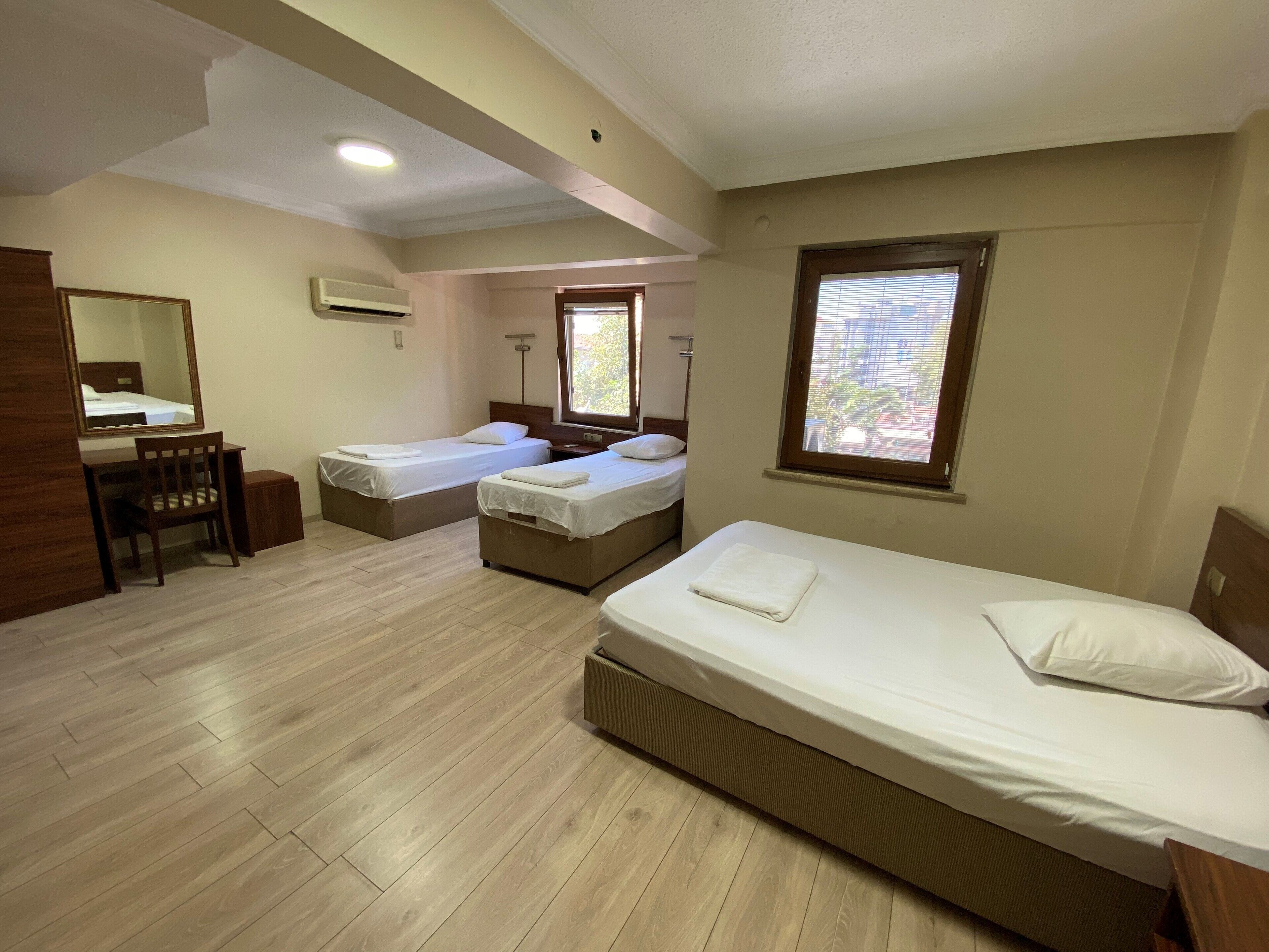 A room with two single beds, a desk with a chair, a large mirror, and a window with blinds.