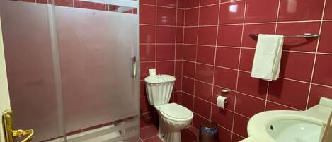 Un baño con paredes de baldosas rojas, un inodoro blanco, una ducha con mampara de vidrio y una bañera blanca.