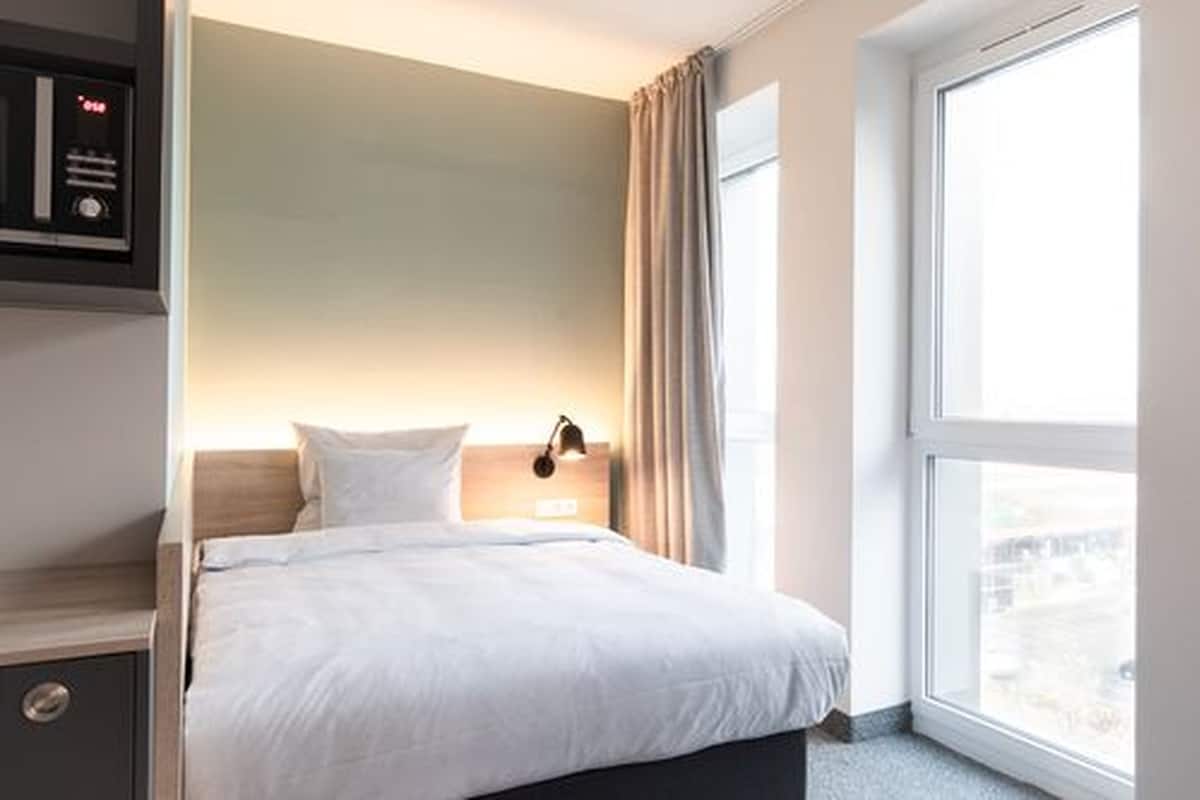 Ein Hotelzimmer mit einem Bett, einem Nachttisch, einer an der Wand befestigten Uhr und einem Fenster mit Vorhängen.