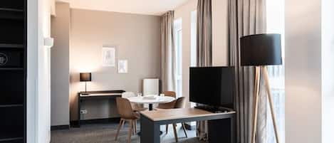 Habitación de hotel con zona de comedor, televisión, lámpara y ventana con cortinas.
