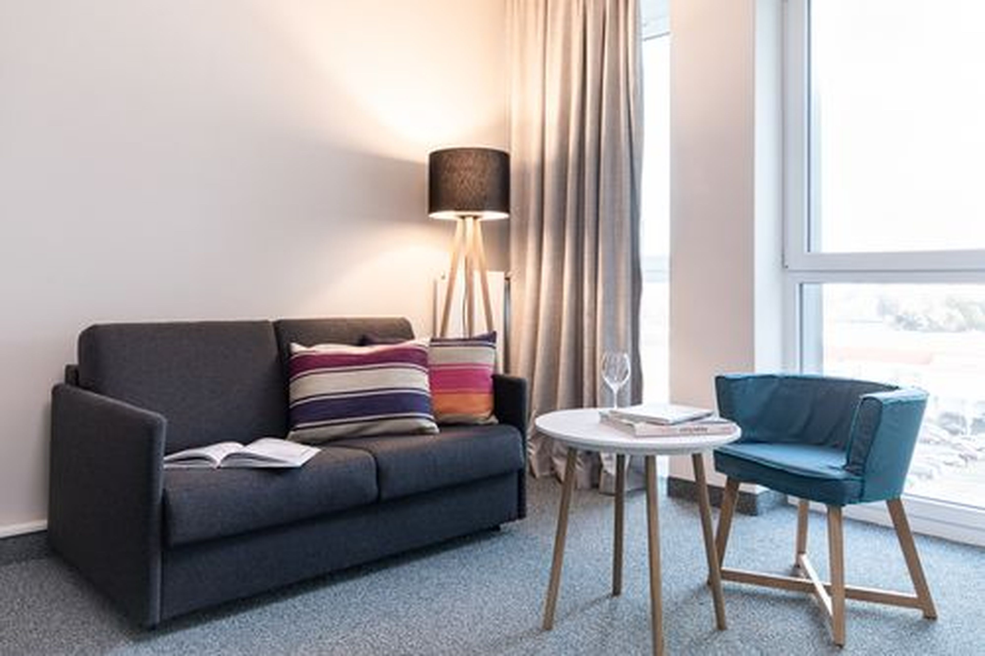 Een moderne woonkamer met een donkergrijze bank, een kleurrijk gestreept kussen, een klein rond tafeltje met een boek en een glas, en een blauwe stoel.