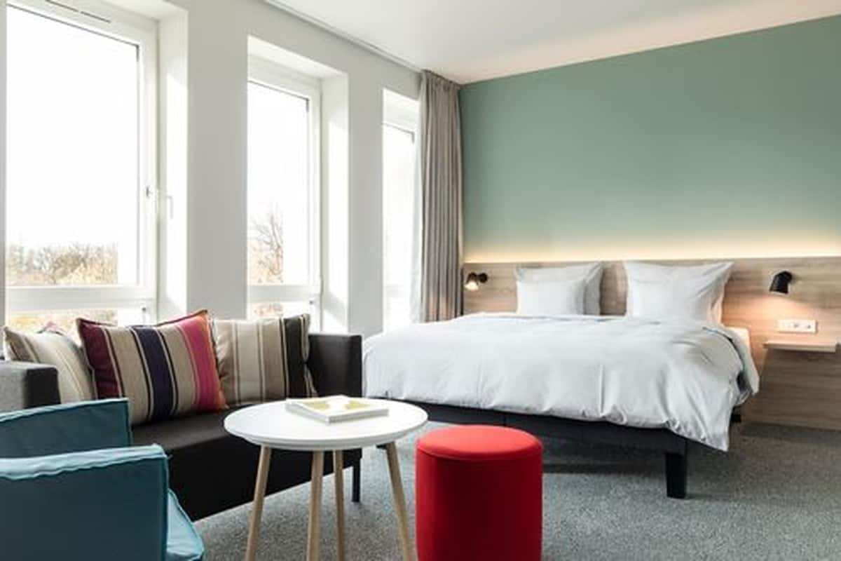 Ein modernes Hotelzimmer mit einem großen Bett, einer Couch, einem kleinen Tisch und einem roten Hocker.