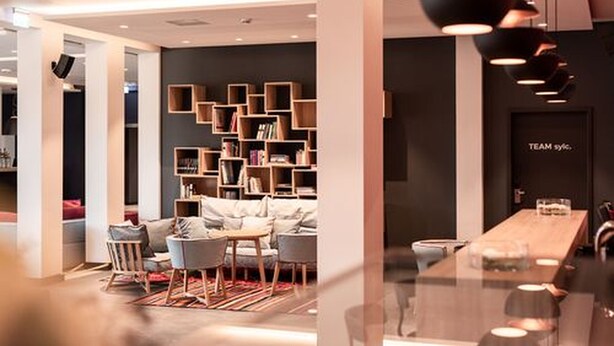 A modern interior with a seating area, a long table, and a bookshelf. - sylc. Apartmenthotel (Hamburg)