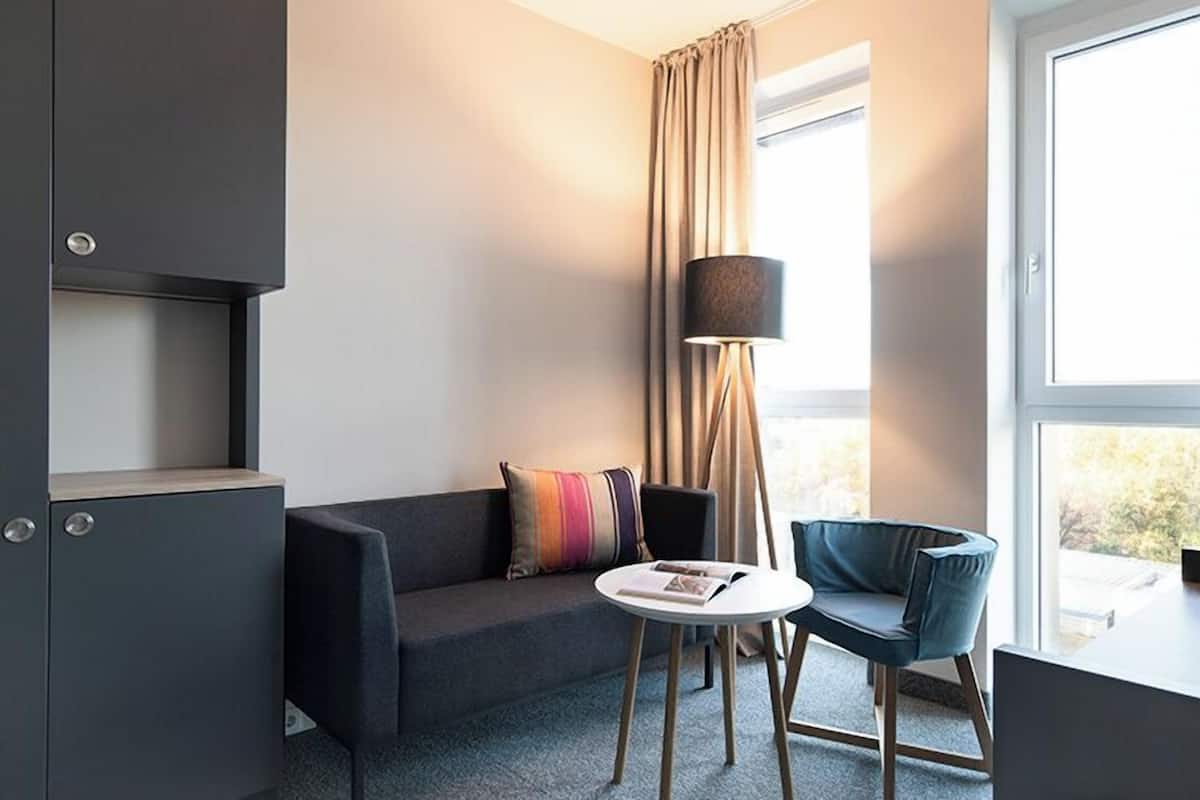 Ein modernes Hotelzimmer mit einem dunkelgrauen Sofa, einem runden weißen Tisch und einem blauen Sessel. Ein Stehlampen und ein Fenster mit Vorhängen sind ebenfalls vorhanden.