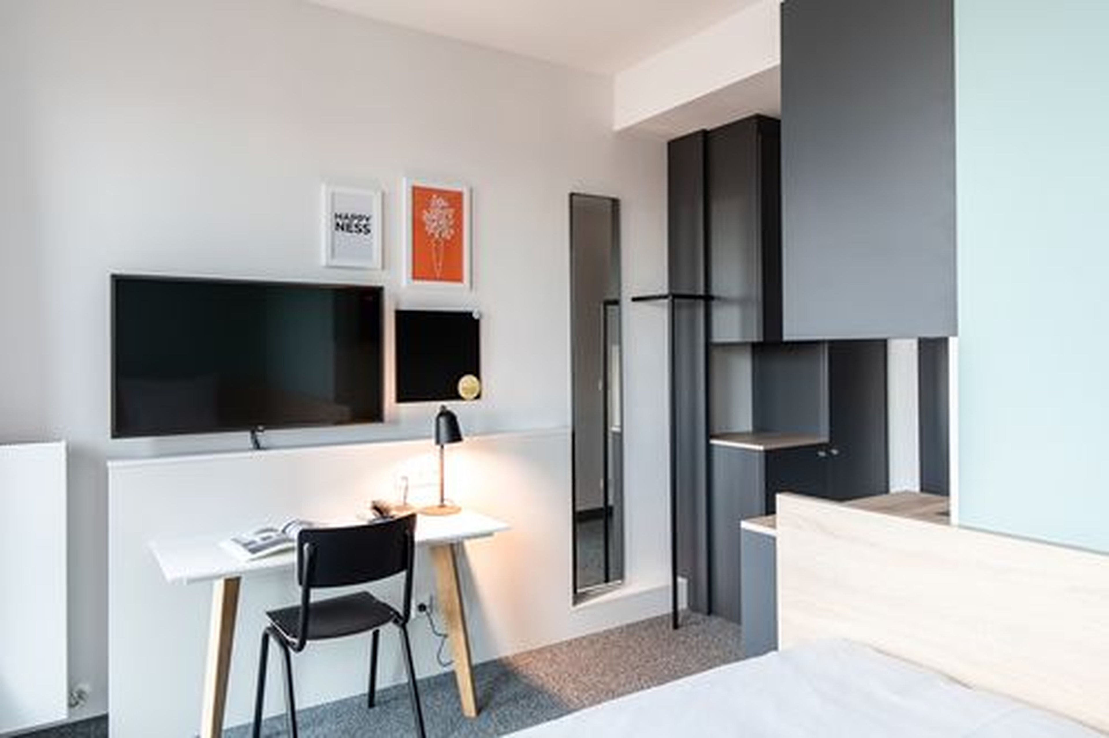 A modern hotel room with a large flat-screen TV, a desk with a chair, a bed, and a large mirror.