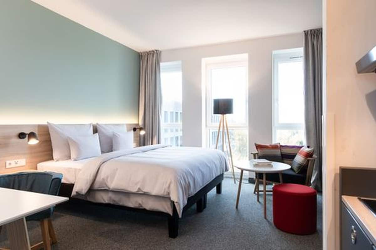 Ein Hotelzimmer mit einem Bett, einem Schreibtisch, einem Sessel, einem kleinen Tisch und einem roten Hocker. Zwei Fenster mit Vorhängen, eine Stehlampe und Blick auf Gebäude sind zu sehen.