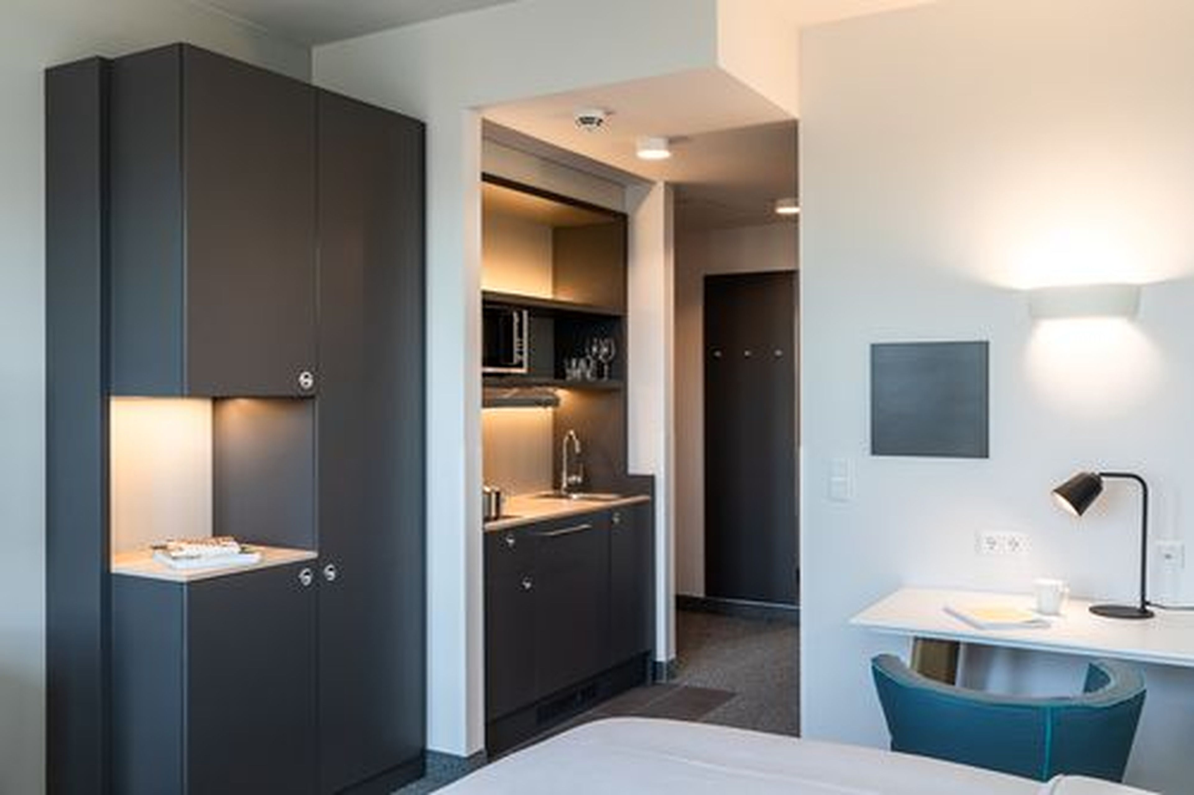 A modern hotel room with a built-in kitchen, a desk with a chair, and a bed.