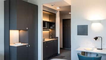 A modern hotel room with a built-in kitchen, a desk with a chair, and a bed.