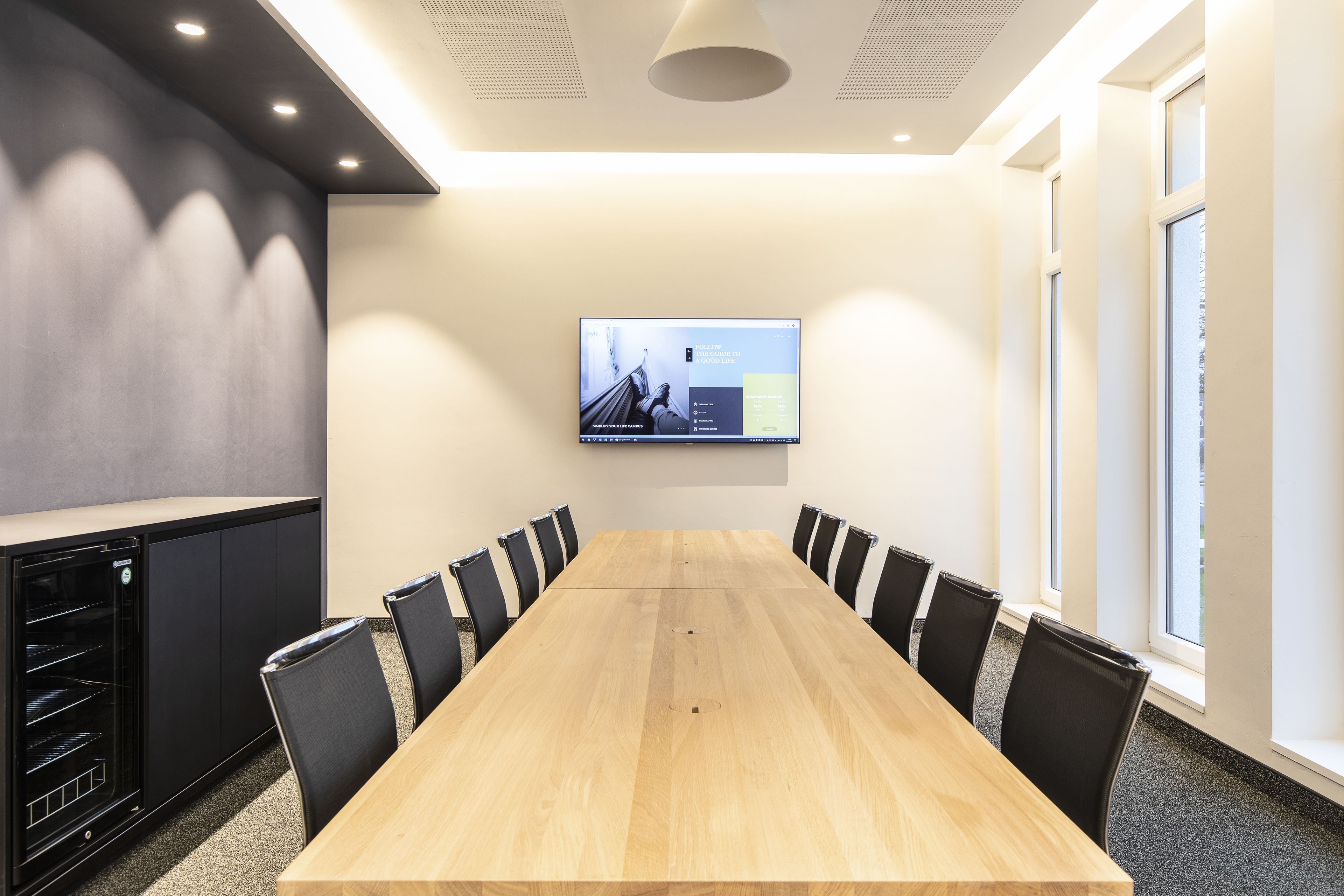 Ein Konferenzraum mit einem langen Tisch, schwarzen Stühlen, einer Wandleinwand und einer Küchenzeile an der Seite.