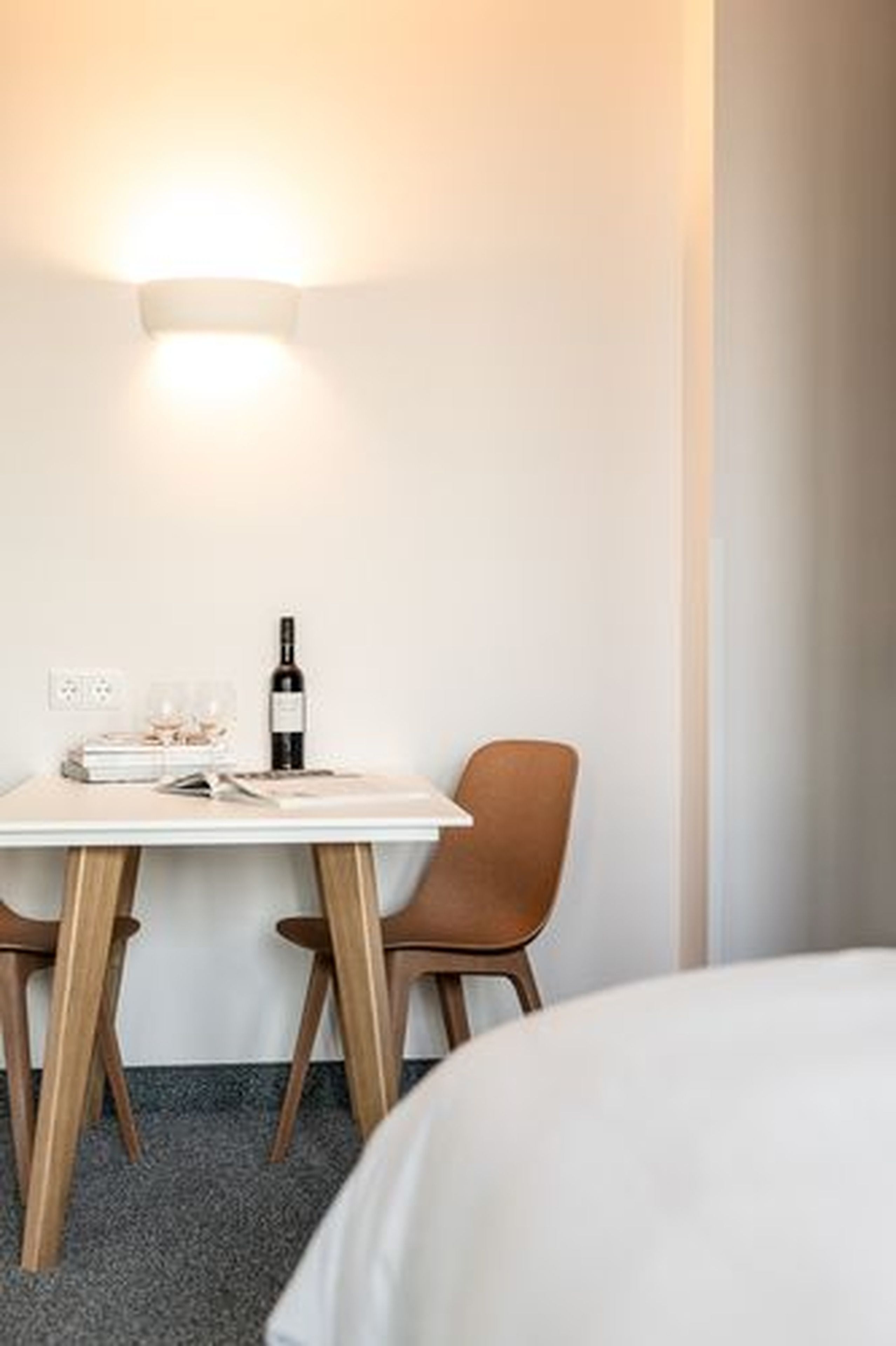 A hotel room with a white bed, a small table with a bottle of wine, a chair, and a wall-mounted light fixture.