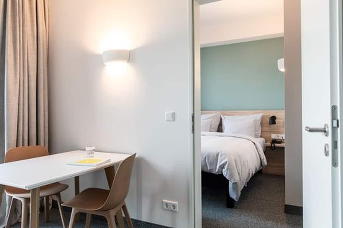 Ein Hotelzimmer mit einem Bett, einem Nachttisch, einem Sessel und einem Tisch mit einem Glas Wasser.