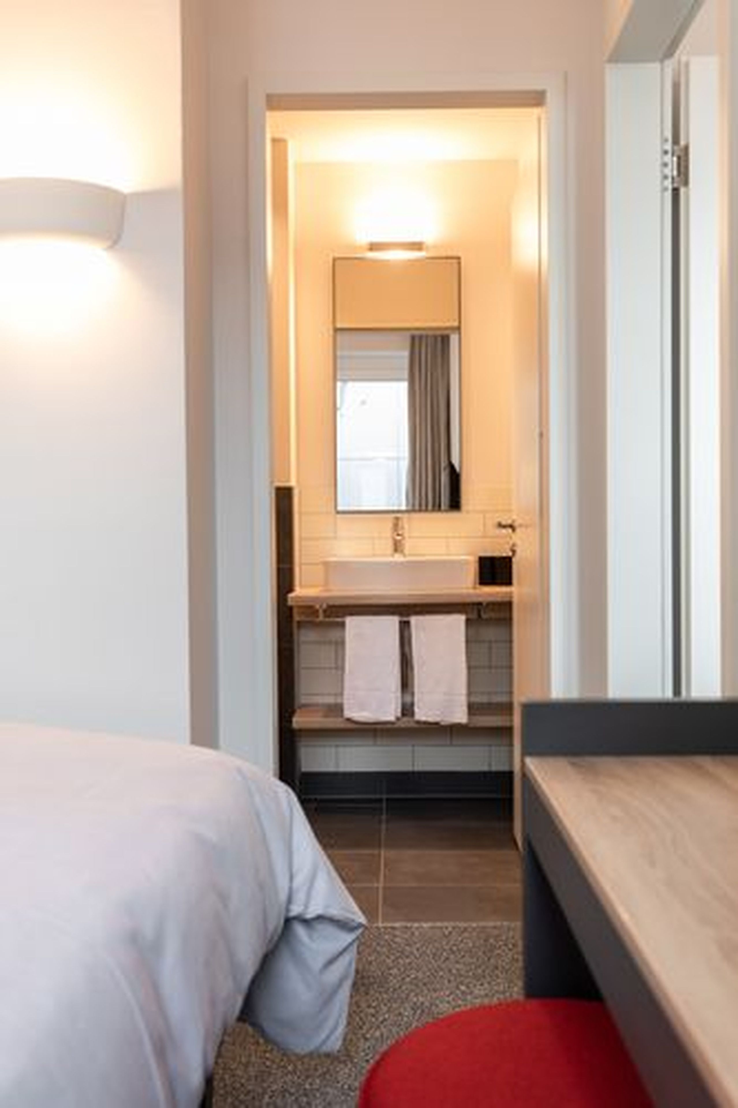 Una habitación de hotel con cama, un banco de madera, una mesita, un lavabo y un espejo.