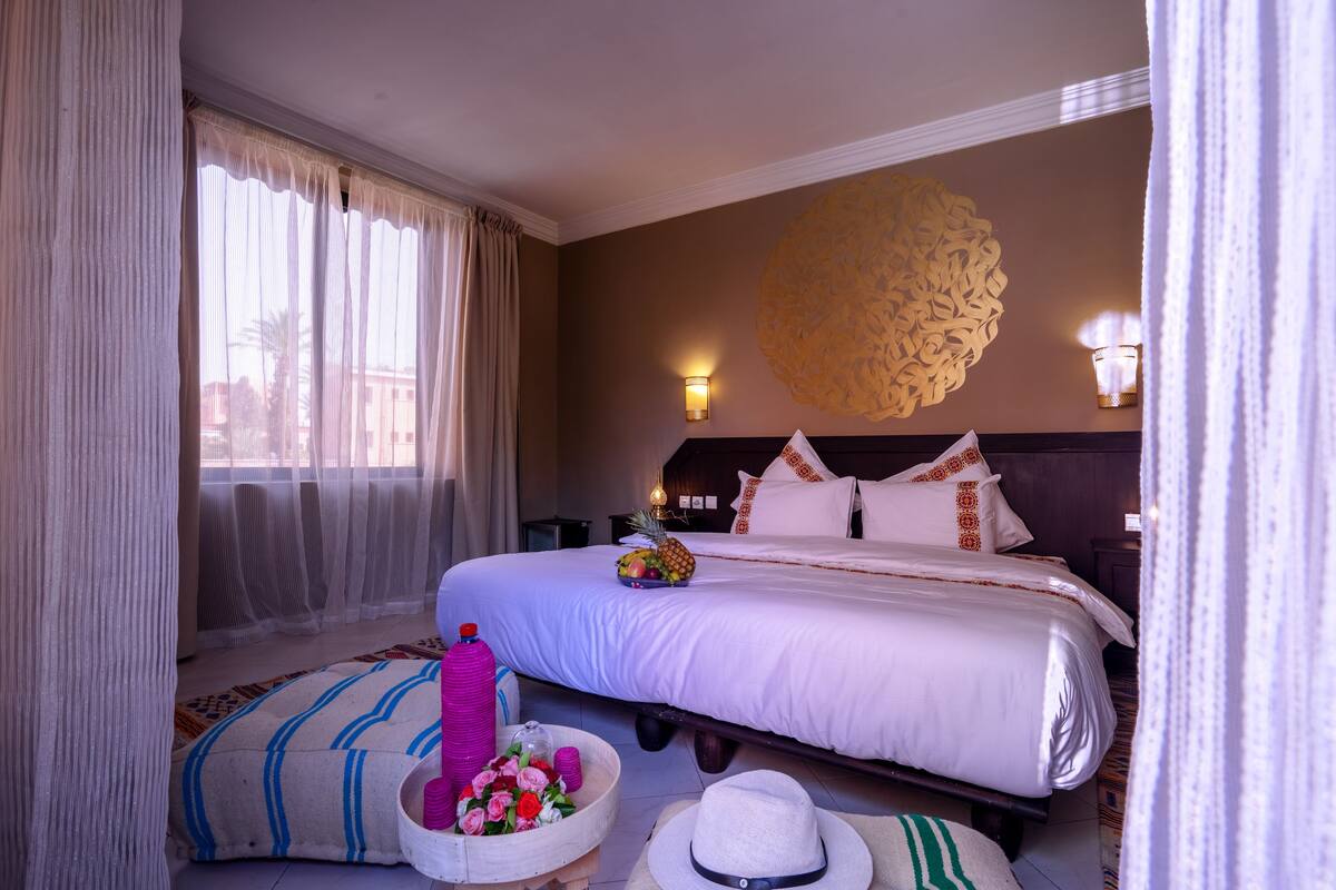 A hotel room with a large bed, a decorative wall piece, and a view of palm trees through sheer curtains.