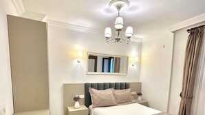 A bedroom with a bed, bedside tables, a mirror, a chandelier, and a window with curtains. - Evren Butik Otel (Bozcaada)