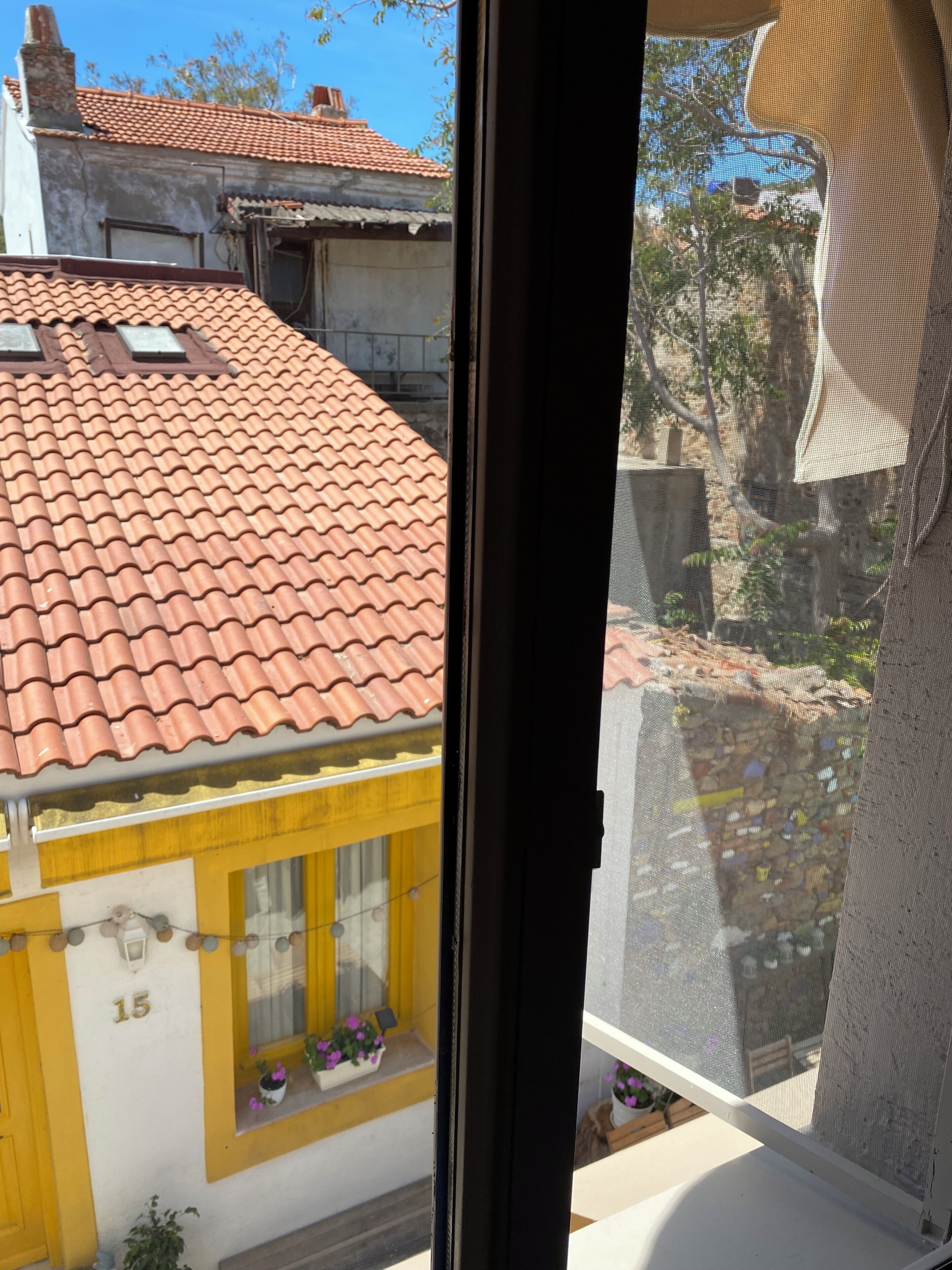A view through a window of a building with a tiled roof, a yellow door, and a window with potted plants.