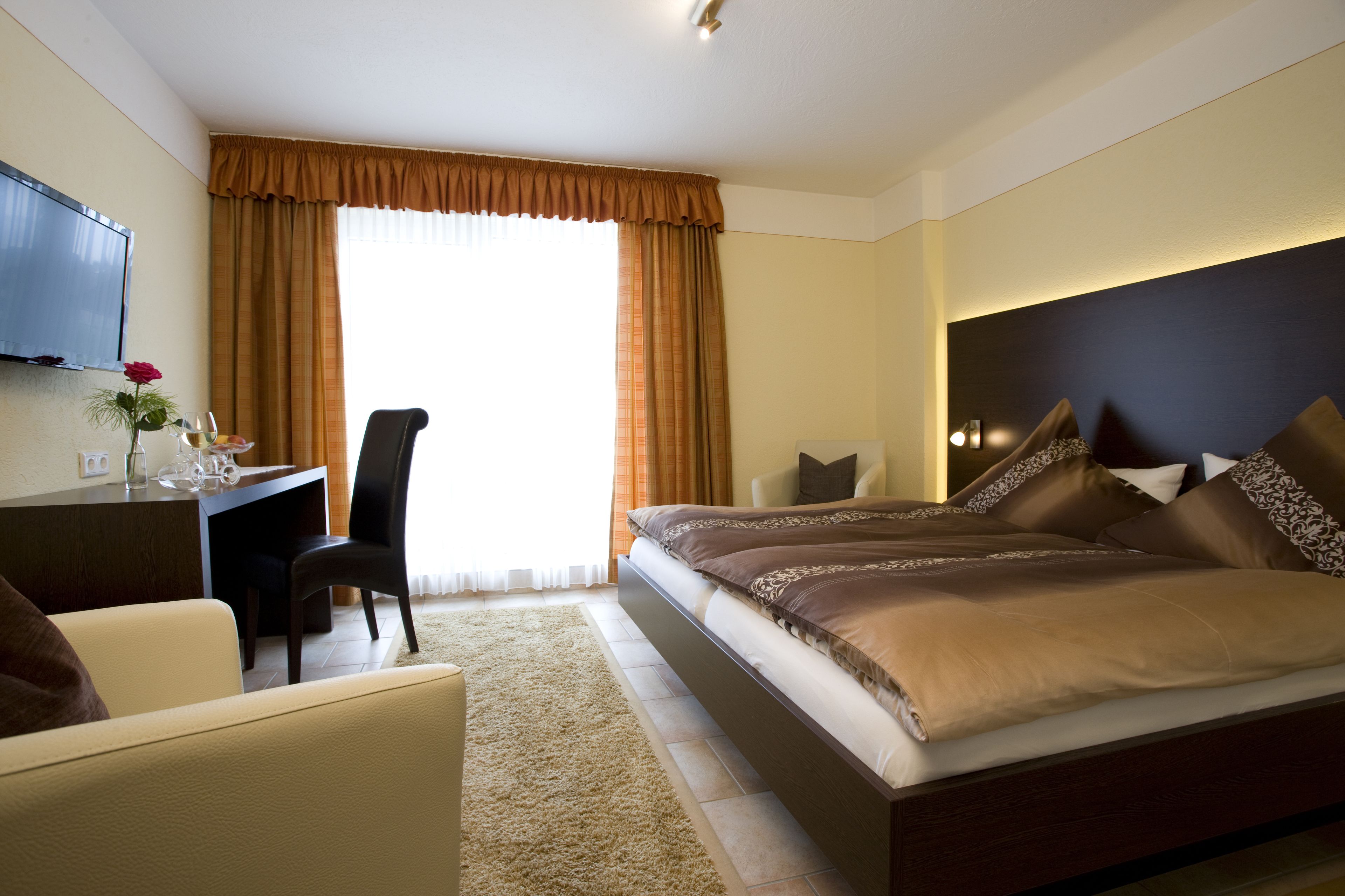 Ein Hotelzimmer mit einem großen Bett, einem Schreibtisch mit Stuhl, einem an der Wand montierten Fernseher und einem Fenster mit Vorhängen.