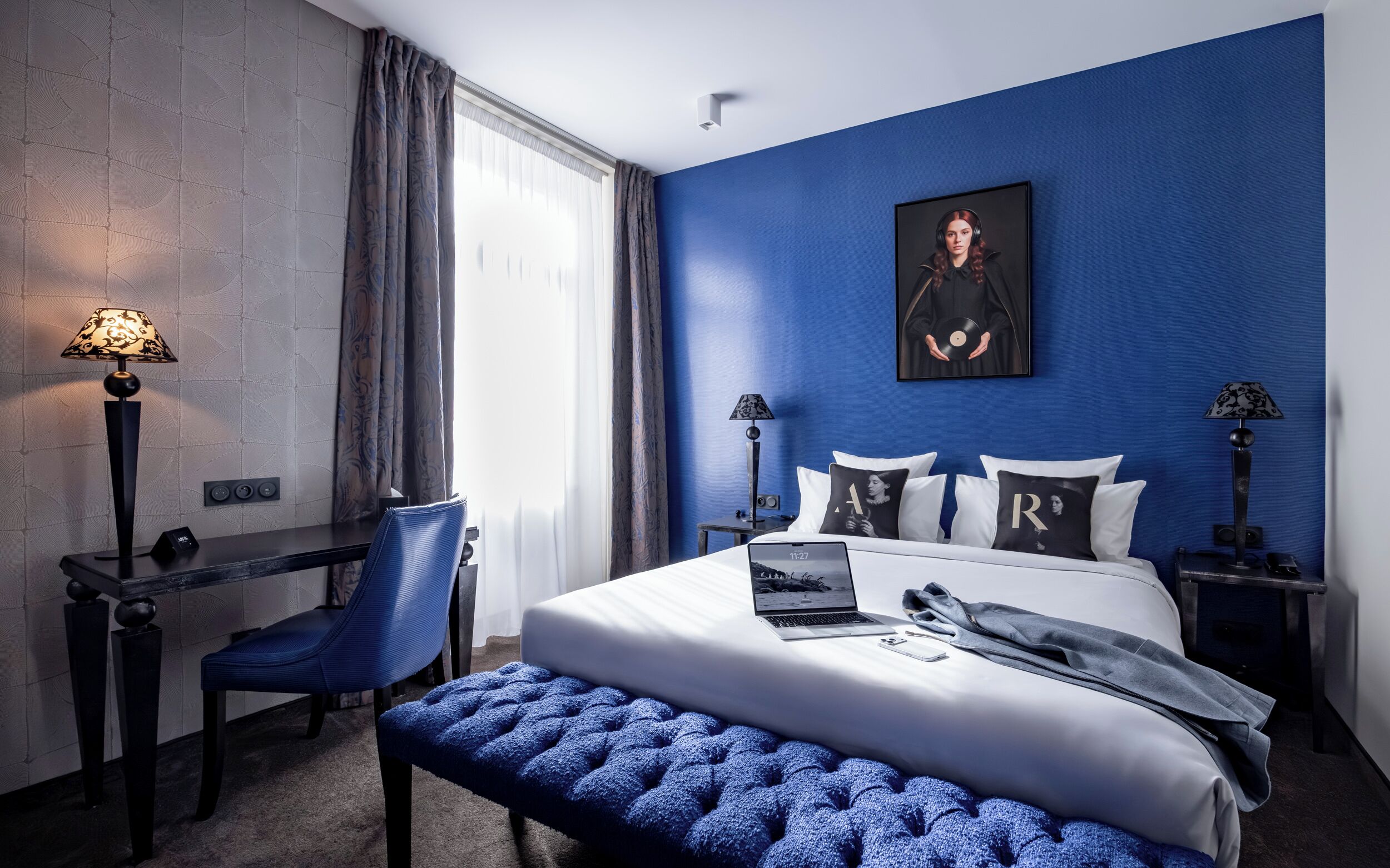 Una habitación de hotel con cama, escritorio, silla, banco y un cuadro en la pared.