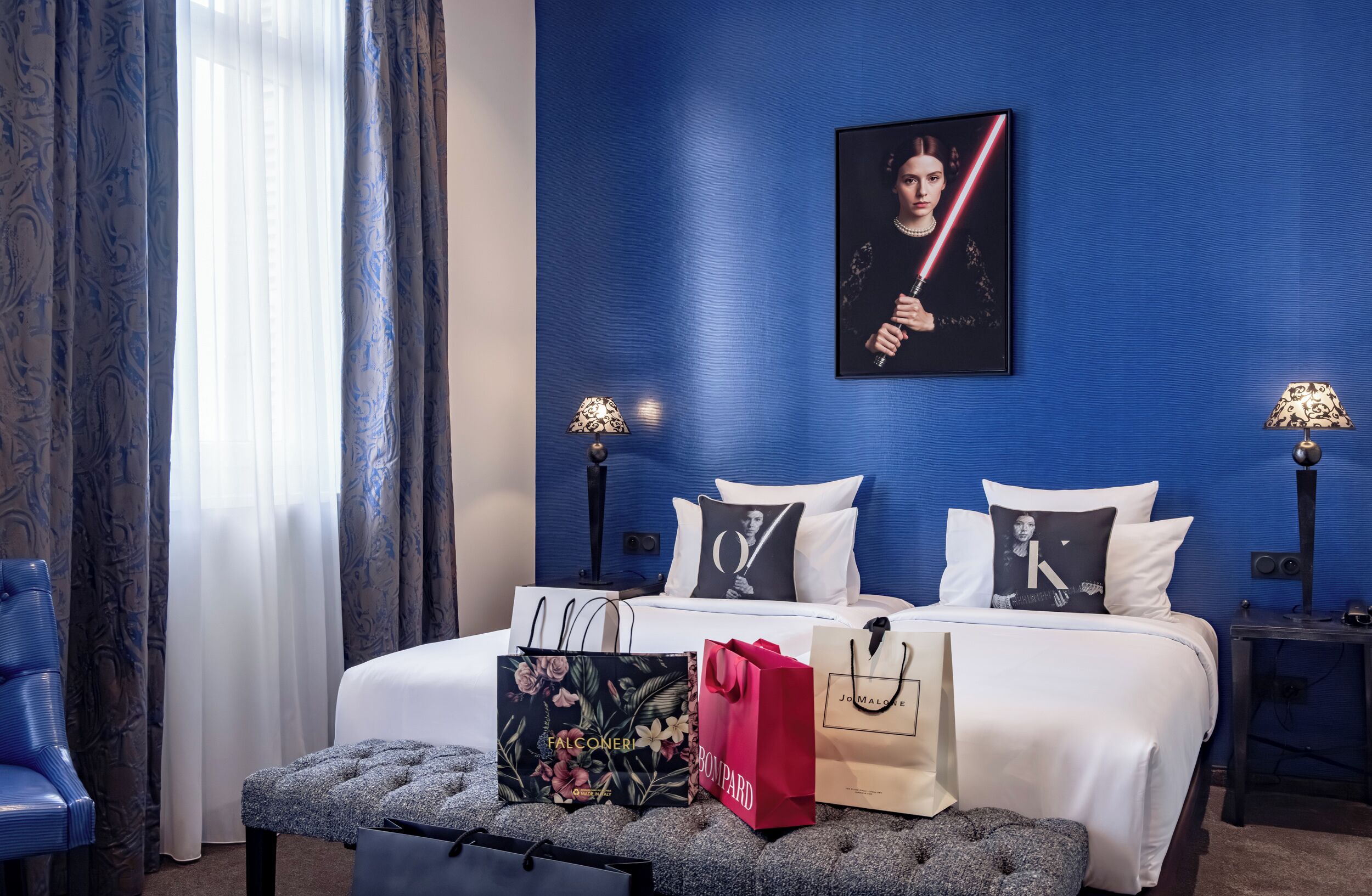 A hotel room with a blue wall, a bed with white bedding, two bedside tables with lamps, a bench with shopping bags, and a poster on the wall.