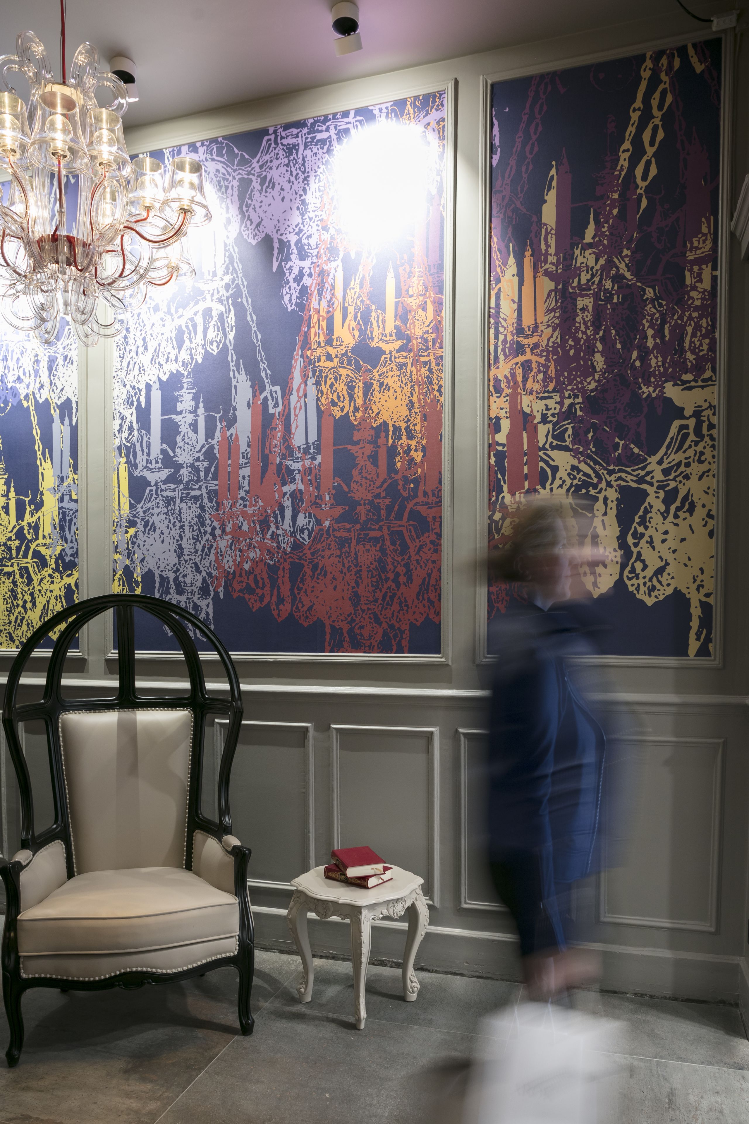 A room with a chandelier, a chair, a small table with a book, and a large abstract painting on the wall.