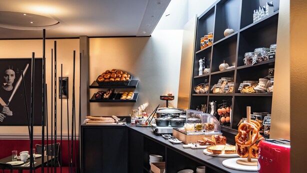 A breakfast bar with pastries, bread, and a red toaster. There are shelves with jars and containers, and a wall with a framed picture. - Hotel Arok (Strasbourg)