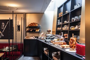 A breakfast bar with pastries, bread, and a red toaster. There are shelves with jars and containers, and a wall with a framed picture. - Hotel Arok (Strasbourg)