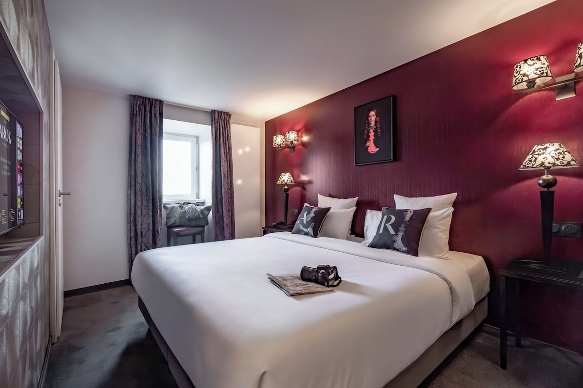 Hotellihuone, jossa on suuri sänky, työpöytä, tuoli, ikkuna verhoilla ja seinään kiinnitetyt lamput.
