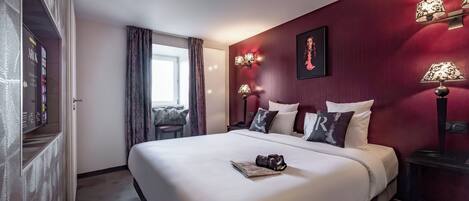 A hotel room with a large bed, a desk, a chair, a window with curtains, and wall-mounted lamps.