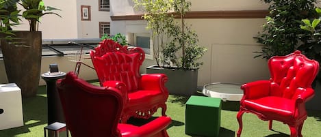 An outdoor seating area with red chairs and a small table, surrounded by potted plants and a green box.