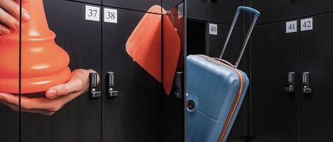A hotel room with black lockers numbered 22 to 42, a blue suitcase with a handle, and an orange object resembling a chess piece.