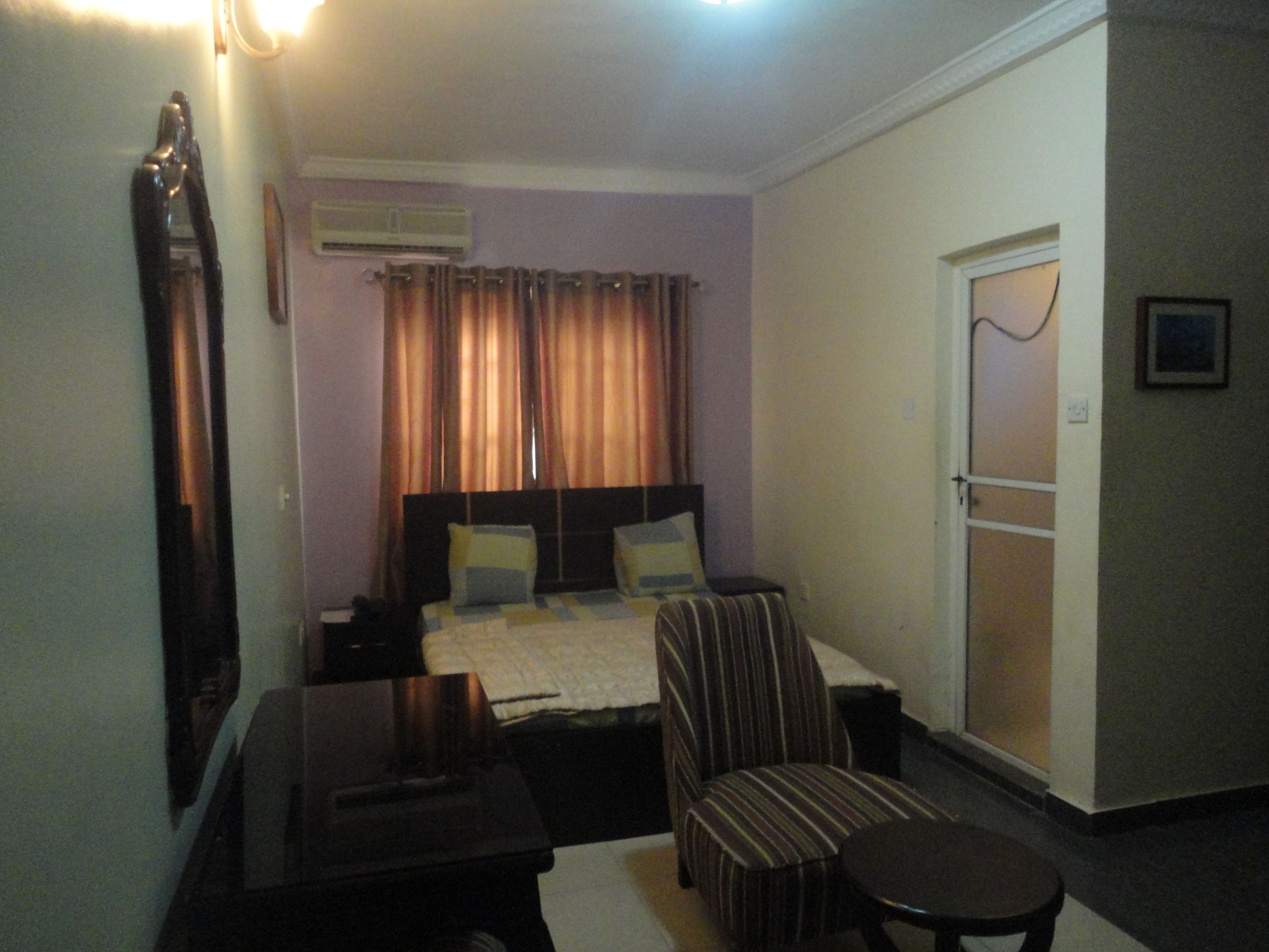 Une chambre d’hôtel comprenant un lit, une chaise, une petite table, des rideaux, un appareil de climatisation et une porte.