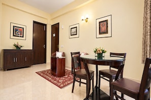 A dining area with a round glass table, four chairs, a red patterned rug, a dark wooden cabinet, and two framed artworks on the wall.