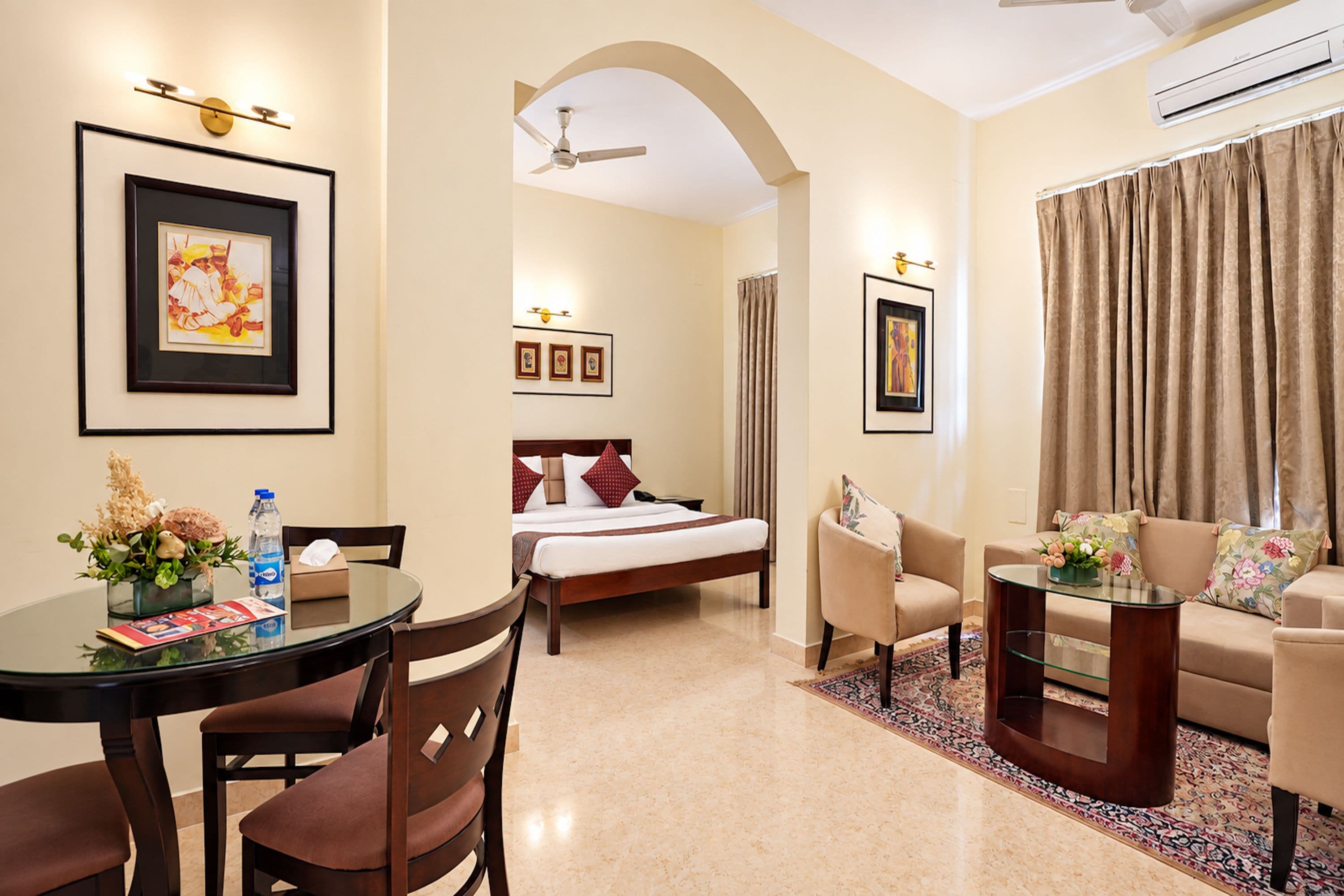 A hotel room with a bed, a sitting area with a sofa and chairs, a dining table with chairs, and a small table with a vase of flowers.