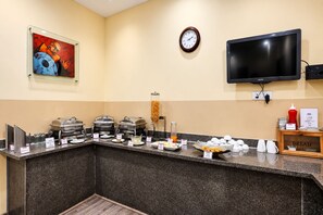 Zona de desayuno tipo buffet con diferentes estaciones de comida, un reloj y un televisor en la pared.