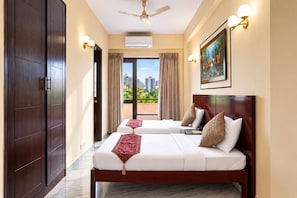 A hotel room with two beds, a ceiling fan, an air conditioning unit, a painting on the wall, and a view of the cityscape through the window.