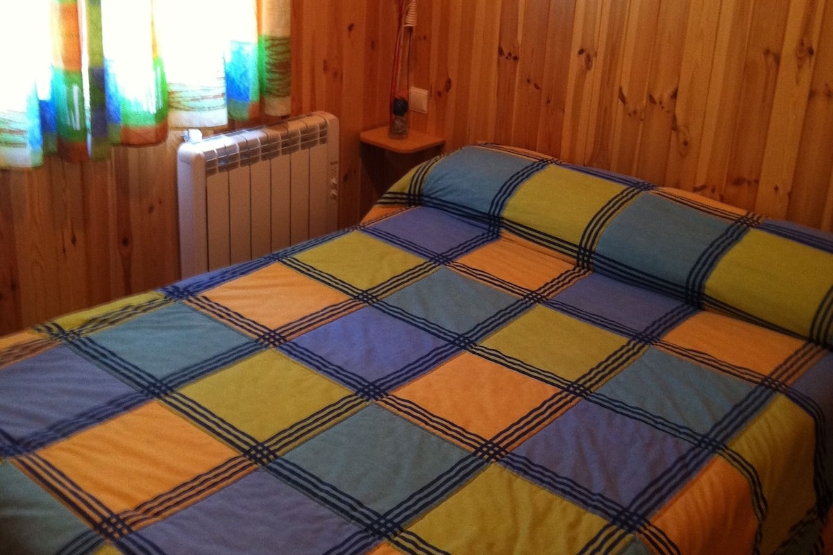 A room with wooden walls, a bed with a colorful quilt, a window with curtains, and a radiator.