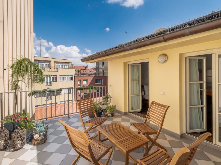 Un balcón con mobiliario de madera y plantas en macetas, con vistas a un barrio residencial con edificios.
