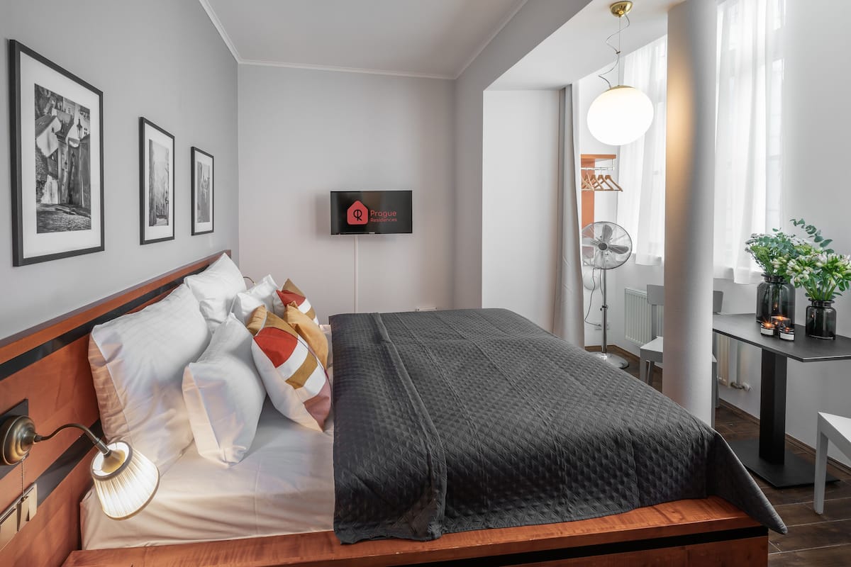 A modern hotel room with a neatly made bed, a flat-screen TV mounted on the wall, and a desk with a fan and a potted plant.