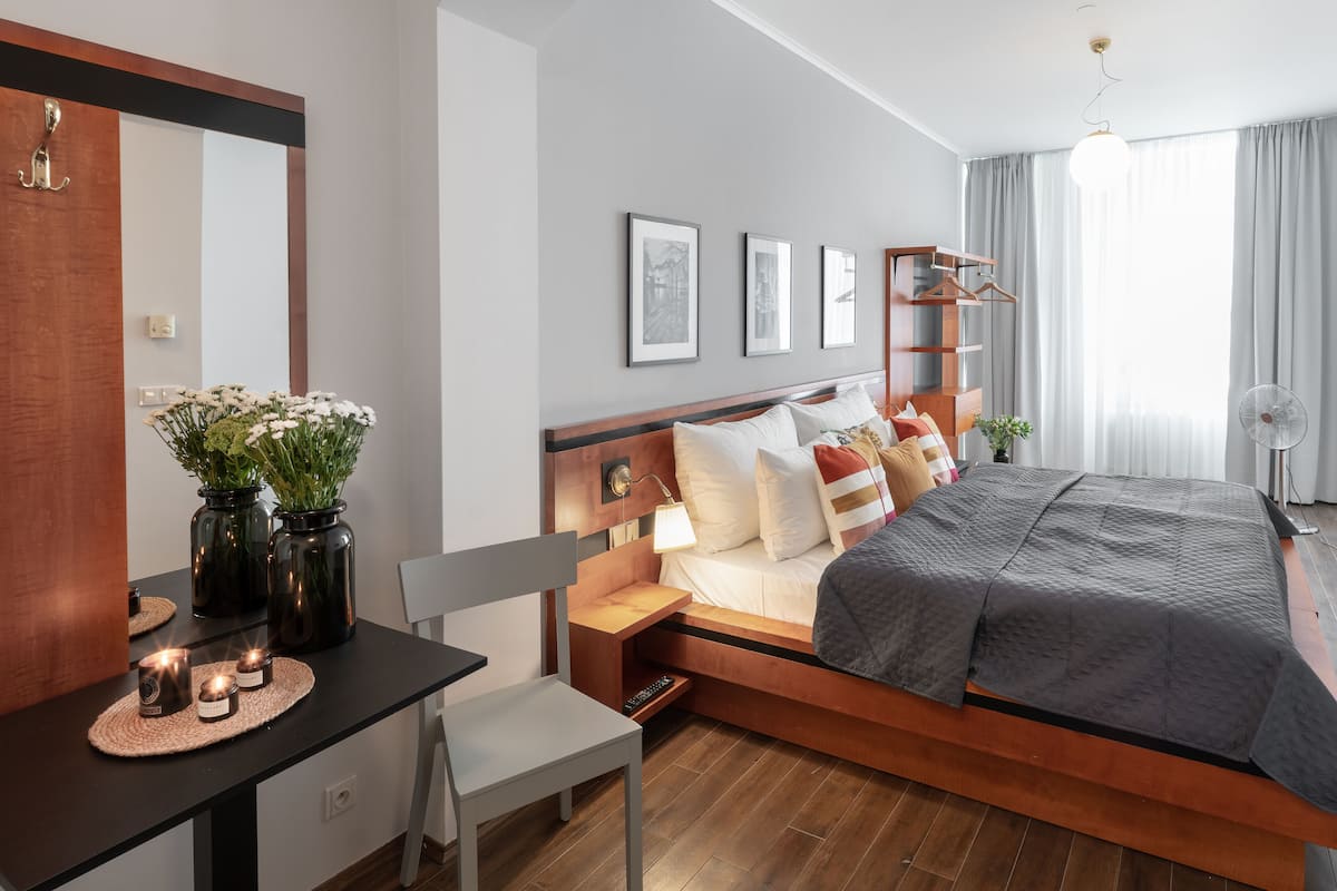 A modern hotel room with a wooden bed, bedside tables, a desk with a chair, and a large mirror.