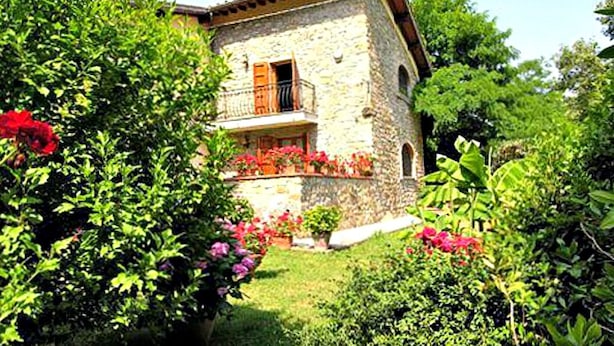 Une maison en pierre dotée d’un balcon, entourée d’une végétation luxuriante et de plantes en fleurs.