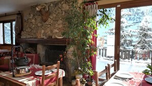 Comedor rústico con chimenea de piedra, mobiliario de madera y vistas a un paisaje nevado a través de amplios ventanales.
