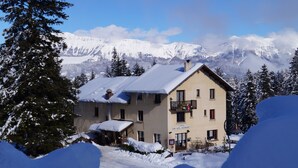 Un edificio cubierto de nieve con un balcón, rodeado de nieve y árboles, con montañas al fondo.