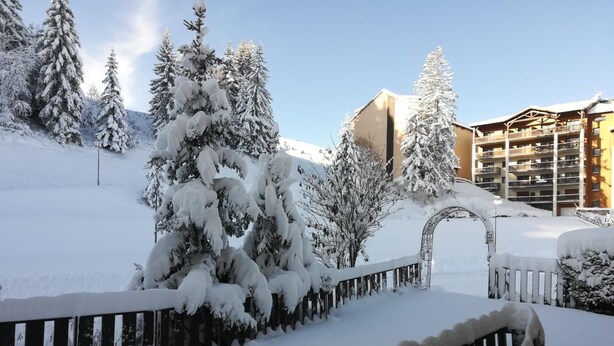 Paisaje nevado con una valla de madera, árboles cargados de nieve y un edificio con balcones.
