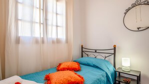 Ein ordentlich bezogenes Bett mit blauer Bettwäsche und orangefarbenen Kissen, ein Nachttisch mit Lampe sowie ein Fenster mit transparenten Vorhängen.