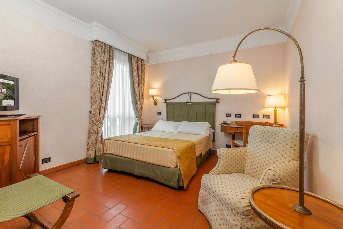 A hotel room with a bed, a chair, a desk, and a lamp.